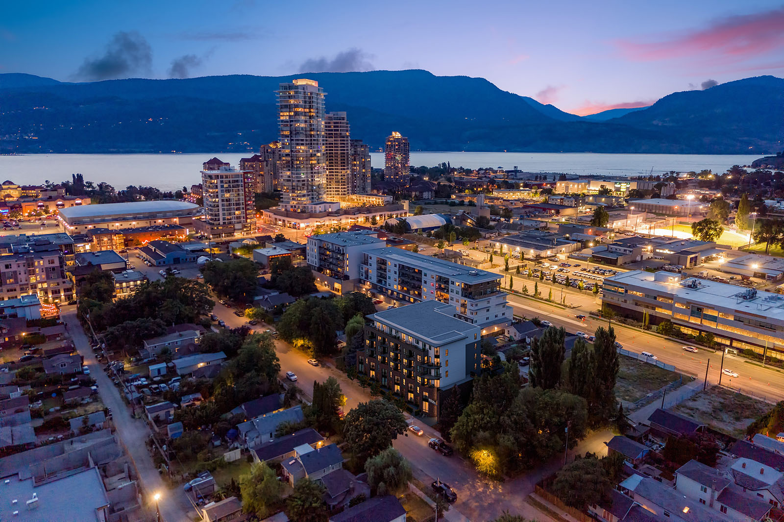Dusk aerial CGI composite of Nolita within downtown Kelowna, with city lights, Prospera Place, Okanagan Lake and mountains beyond.